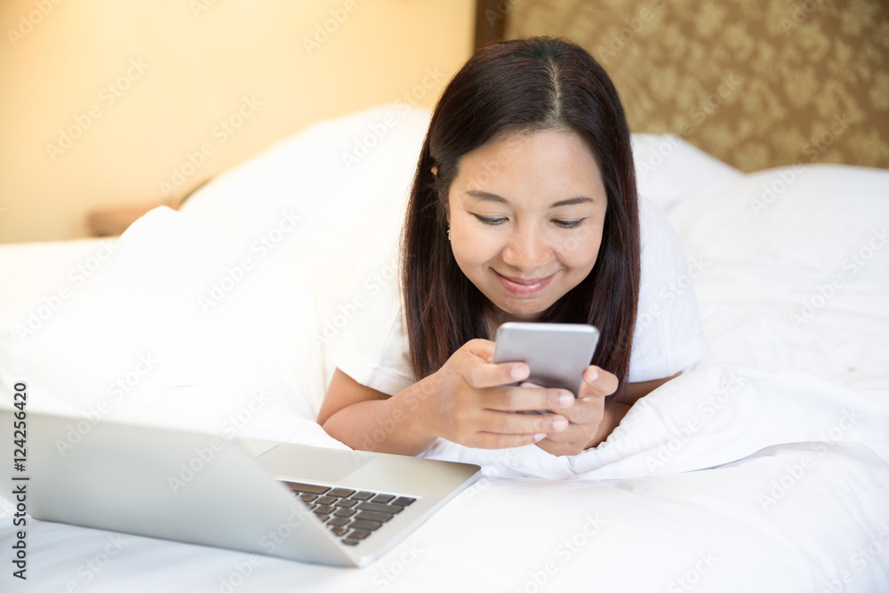 Asian casual woman working on a laptop laying on the white bed, Asian woman looking smartphone on bed Asian casual woman working on a laptop laying on the white bed, Asian woman looking smartphone on bed