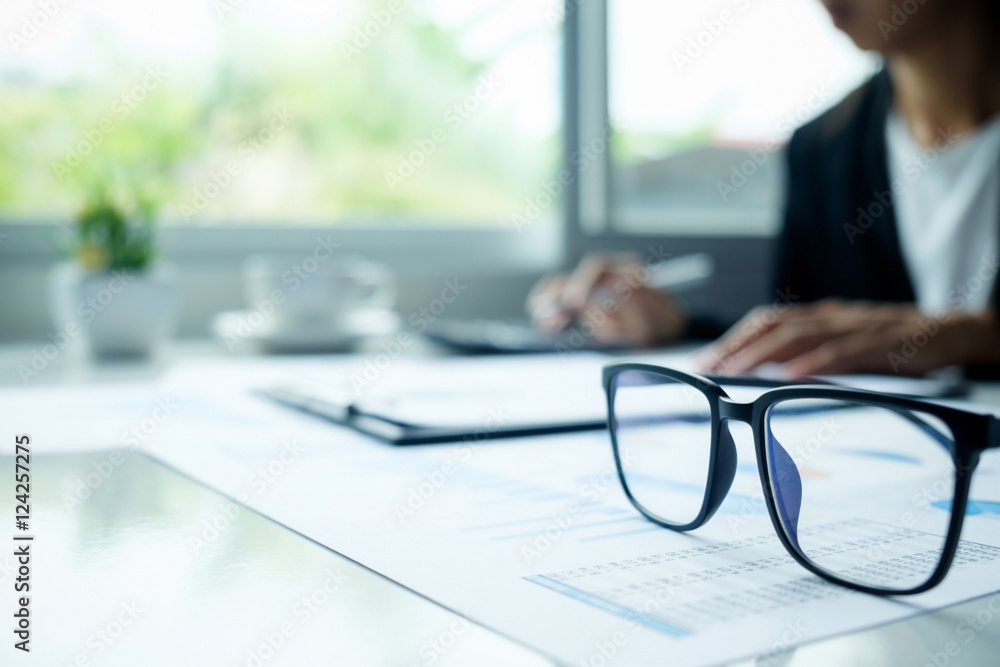 Office desk table with eyeglasses focus and asian business woman working background. Vintage tone Retro filter effect,soft focus,low light.(selective focus). Office desk table with eyeglasses focus and asian business woman working background. Vintage tone Retro filter effect,soft focus,low light.(selective focus).