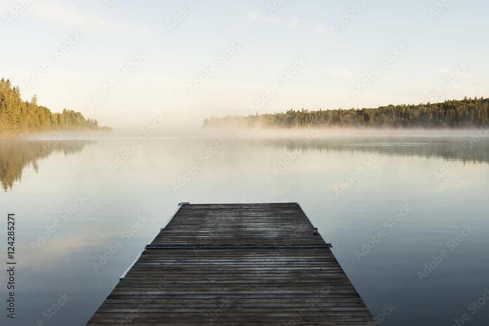 A wooden dock leading out to a misty tranquil lake in Riding Mountain National Park; Wasagaming, Manitoba, Canada A wooden dock leading out to a misty tranquil lake in Riding Mountain National Park; Wasagaming, Manitoba, Canada