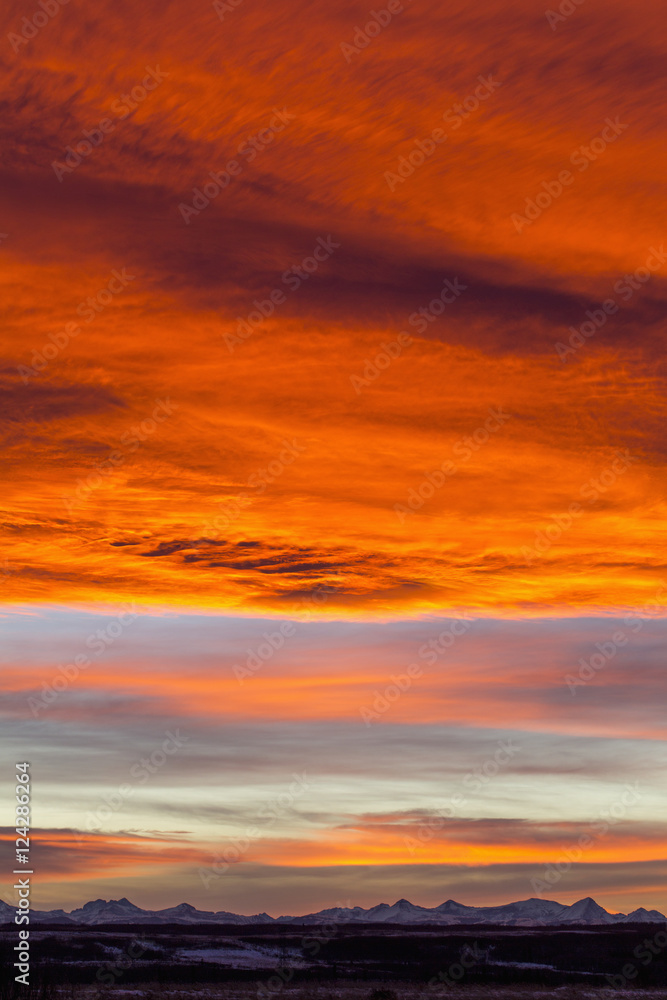 Dramatic fiery sunset with orange and red clouds and mountains in the distance; Calgary, Alberta, Canada Dramatic fiery sunset with orange and red clouds and mountains in the distance; Calgary, Alberta, Canada