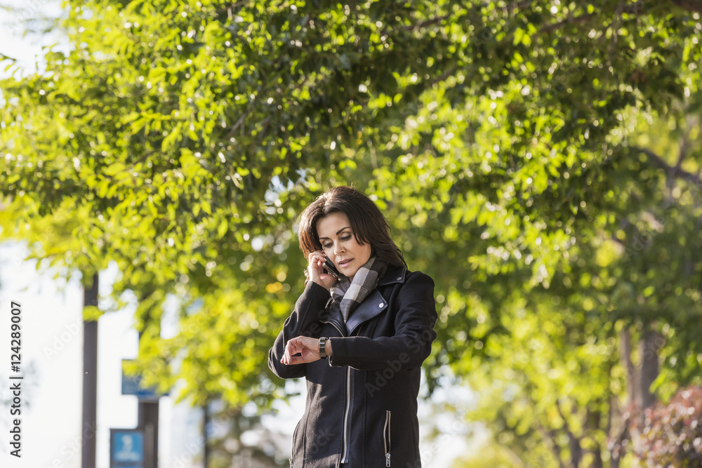 Mature businesswoman talking on her cell phone and checking the time while walking in an urban park outdoors in autumn; Edmonton, Alberta, Canada Mature businesswoman talking on her cell phone and checking the time while walking in an urban park outdoors in autumn; Edmonton, Alberta, Canada