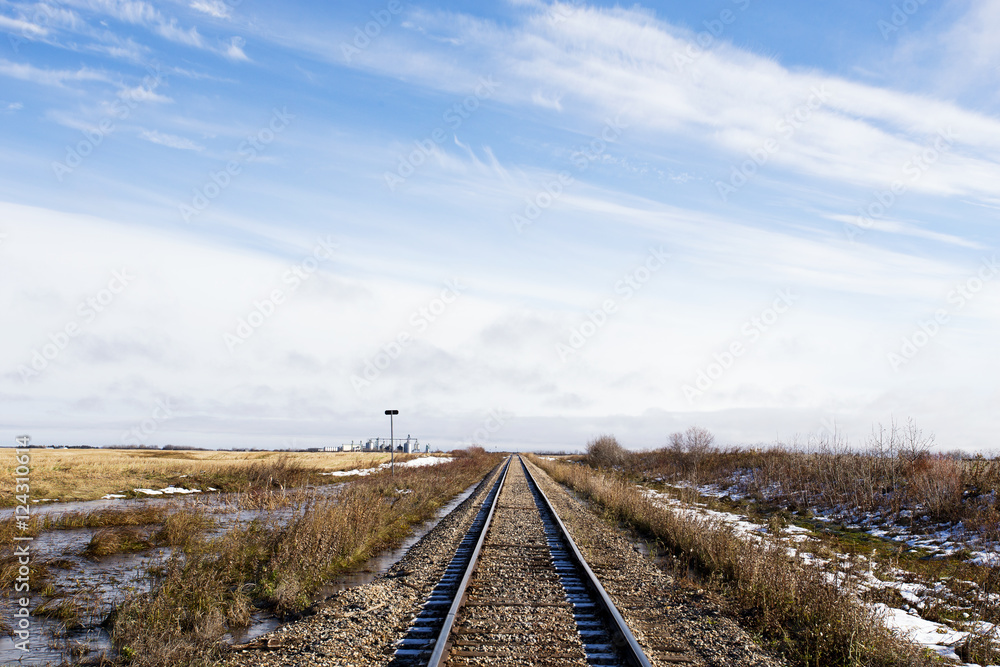 Railway track running straight ahead between fields with a grain terminal in the distance in snow dusted rural saskatchewan landscape