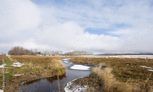 Fototapeta Naklejka Na Ścianę i Meble -  A stream dotted with snow patches winding through a harvested field under cloudy sky in rural landscape
