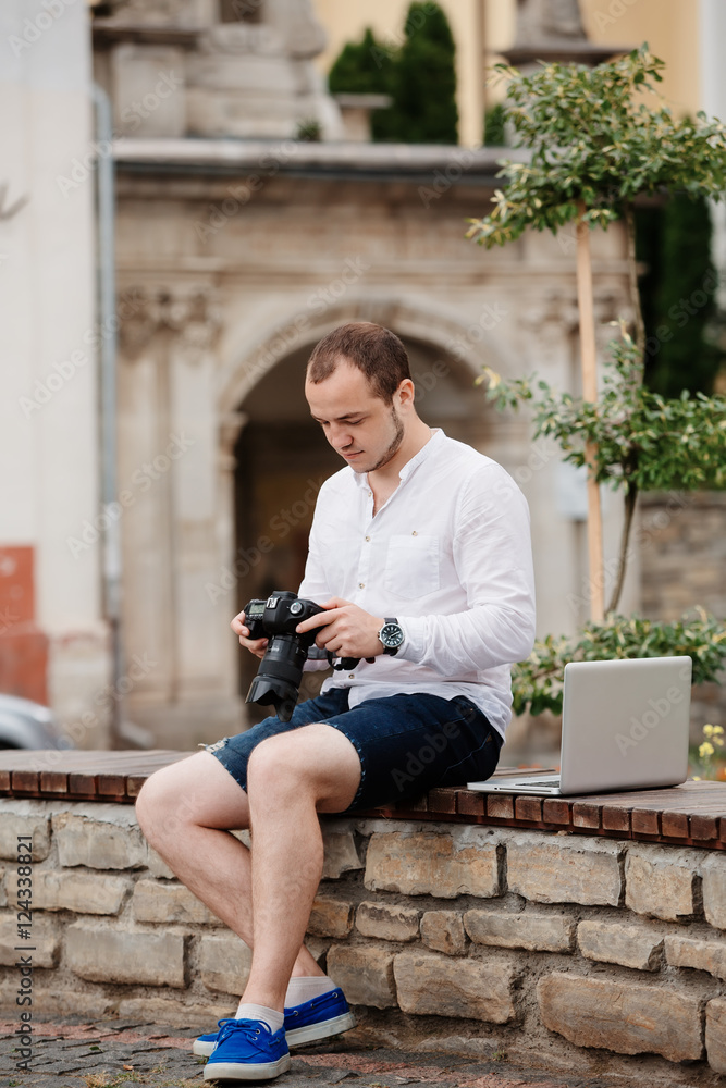 Obraz premium Young photographer with laptopon stone bench andtaking photo on camera