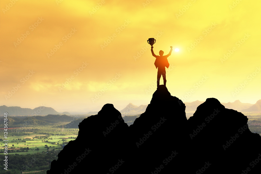 Business success concept.Silhouette of victory businessman hold trophy on top of the mountain with sunset sky as background