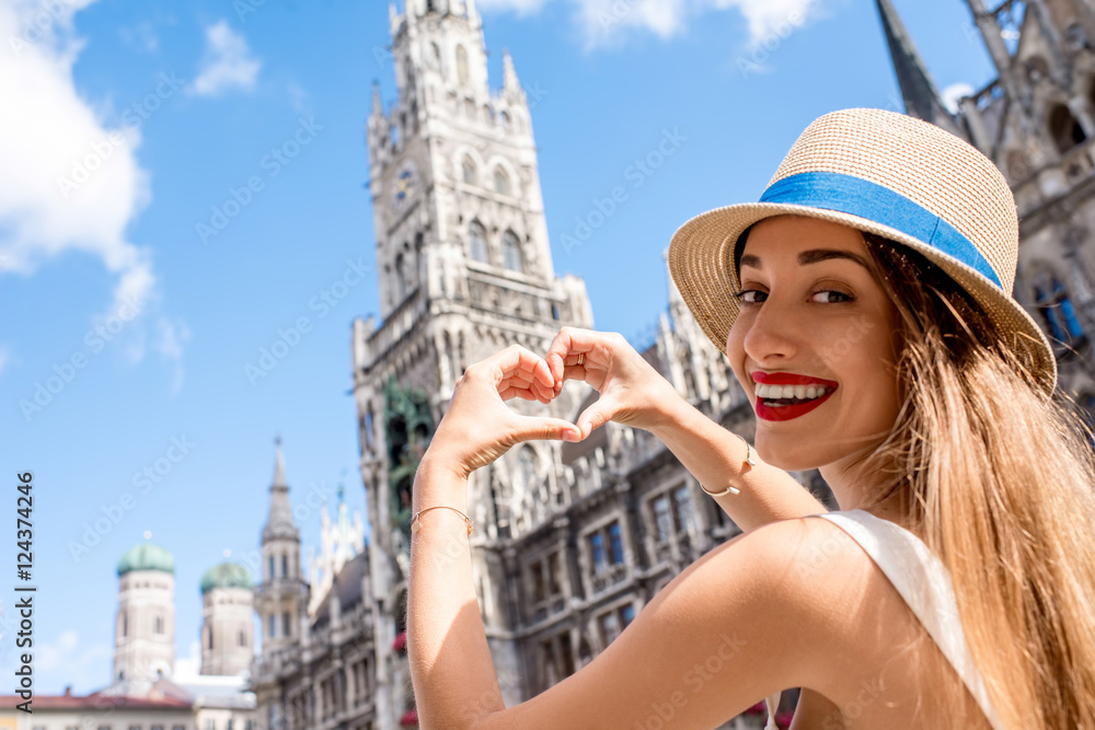 Fototapeta premium Young female tourist making heart shape with hands on the town hall building background in Munich. Having a great vacation in Germany