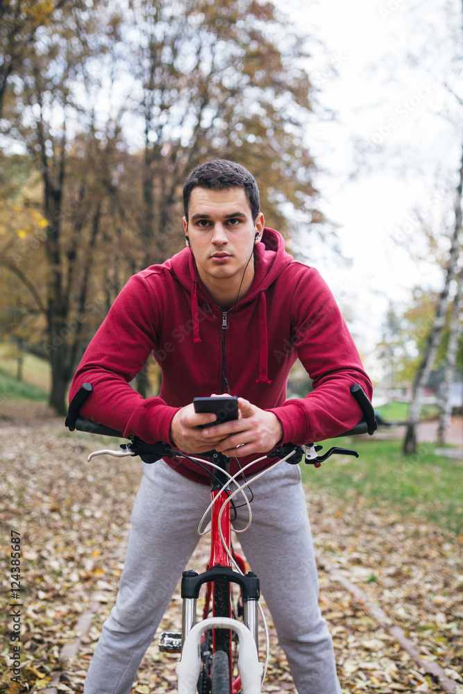 Autumn outdoors. Young serious handsome guy with earphones standing with bicycle in park, holding a cell phone and looking around.