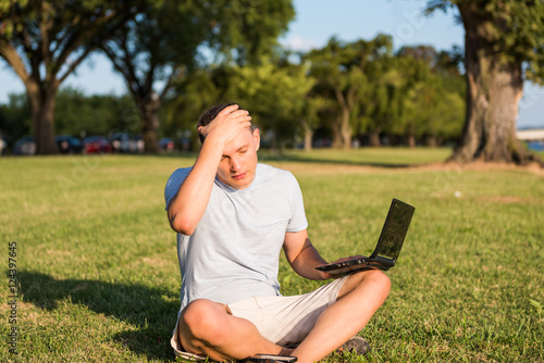 Sad young man with laptop and smartphone, sitting outside in green grass in park placing hand on head due to stress headache and eyes closed looking down photo