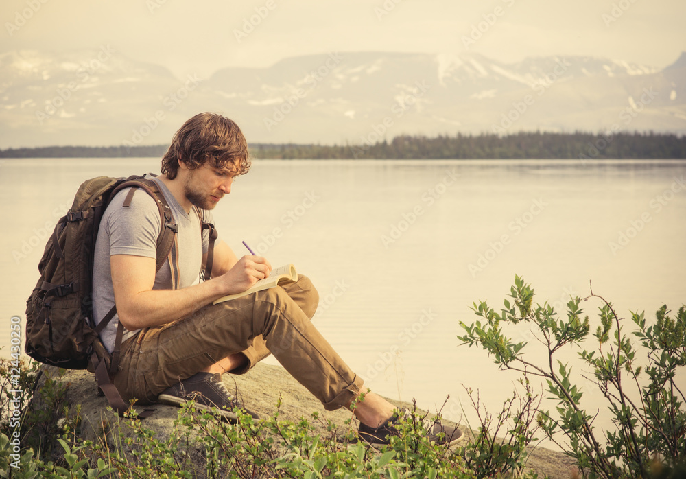 Young Man Traveler with backpack reading book and writing notes outdoor mountains on background Summer vacations and Lifestyle concept