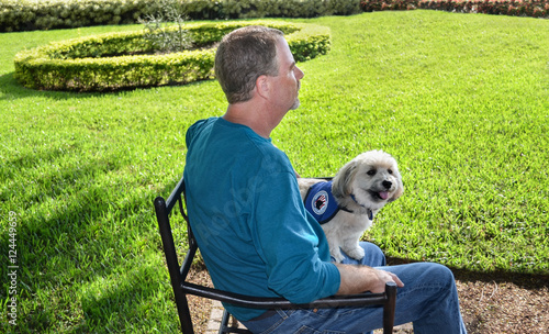 Older gentleman sitting on bench enjoying the sunshine with his Havanese service dog standing on his lap.