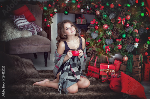 красивая молодая брюнетка сидит рядом с камином около рождественской елки и пьет чай из красной кружки.