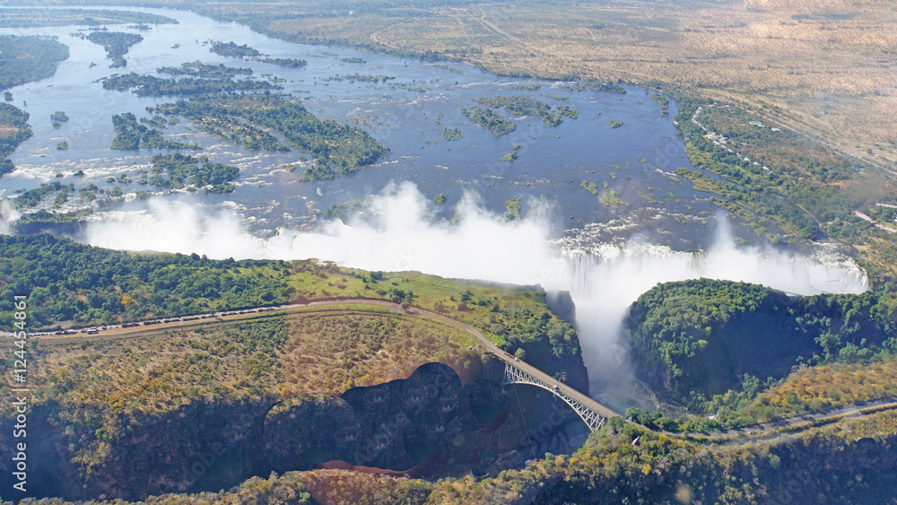 Fototapeta premium Sambesi und Victoriafälle aus der Luft /Blick aus der Luft auf die Victoriafälle, Grenzbrücke zwischen Samia und Simbabwe über einen Canyon, Sambesi mit bewaldeten Inseln