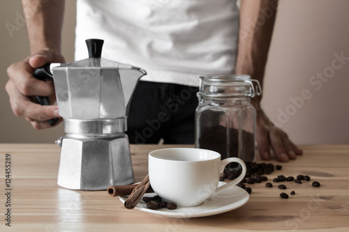 Hands holding coffee pot ready to pour espresso in the white cup