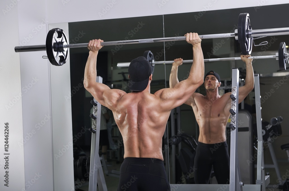 Naklejka premium Handsome powerful athletic man doing barbell shoulder press exercise. Strong bodybuilder with perfect muscles.