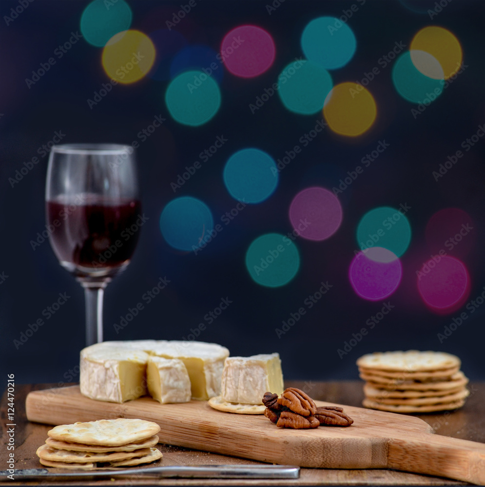 Brie and pecan halves on wooden cheese board, accompanied by crackers and one glass of red wine, against black background, decorated with colorful holiday lights- eating alone