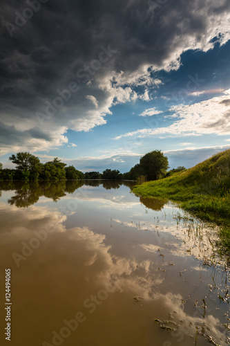Ominous stormy sky over natural flooded river, with bright sun emerging from under the cumulus cloud cover © Calin Tatu