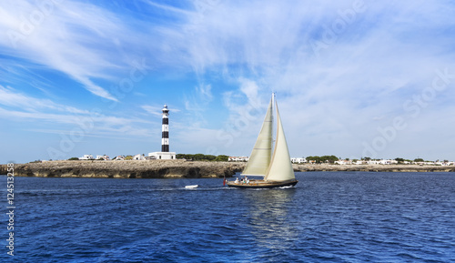Sailboat sailing on mediterranean sea with a lighthouse on foregroud. Menorca Island, Spain photo