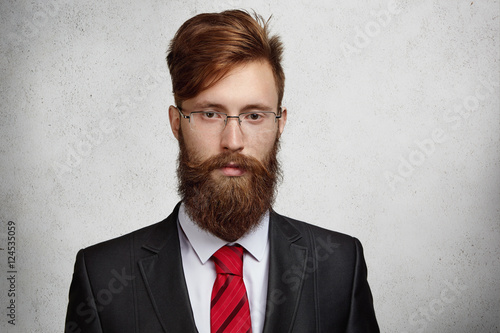 Close up shot of attractive young bearded corporate worker in spectacles and elegant suit on his first working day at big financial company, having focused and confident expression, ready to work hard