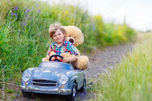 Little kid boy driving big toy car with a bear, outdoors.