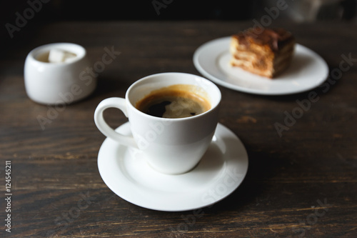 A white cup of black coffee  a piece of cake on the white plate  sugar bowl on the wooden table in a cafe. Selective focus  small depth of fieild.