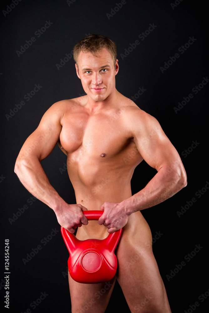 Naklejka premium Totally Naked muscular man covering with red kettlebell, intriguingly smiling