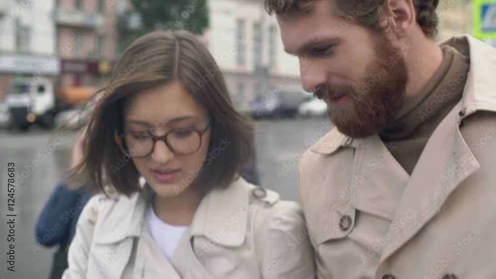 Handheld dolly-like shot with tilt up and down of stylish bearded man and woman in glasses crossing street chatting and looking at tablet, people with gadgets in background Handheld dolly-like shot with tilt up and down of stylish bearded man and woman in glasses crossing street chatting and looking at tablet, people with gadgets in background