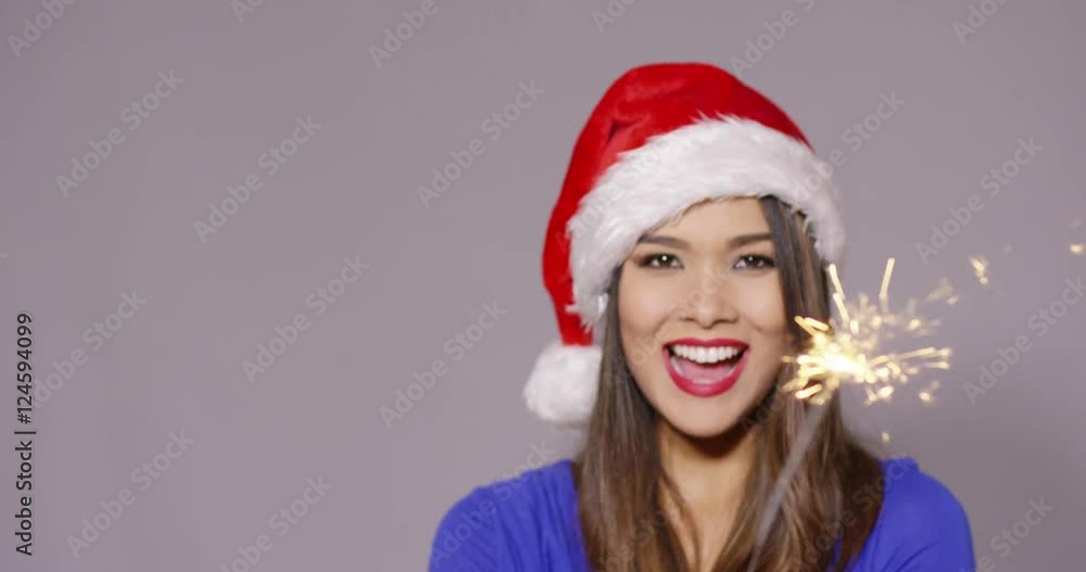Gorgeous sexy young woman in a festive red Santa hat celebrating Christmas with a burning sparkler in her hand and a beaming smile