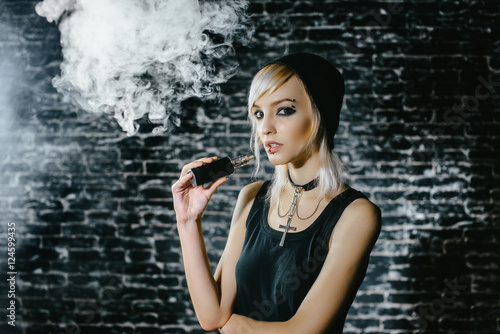 Sexy goth girl smokes electronic cigarette on dark background. The model vaper vaping a vaporizer in the studio.