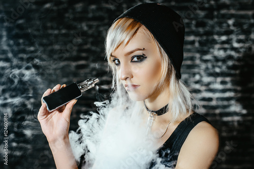 Sexy goth girl smokes electronic cigarette on dark background. The model vaper vaping a vaporizer in the studio.