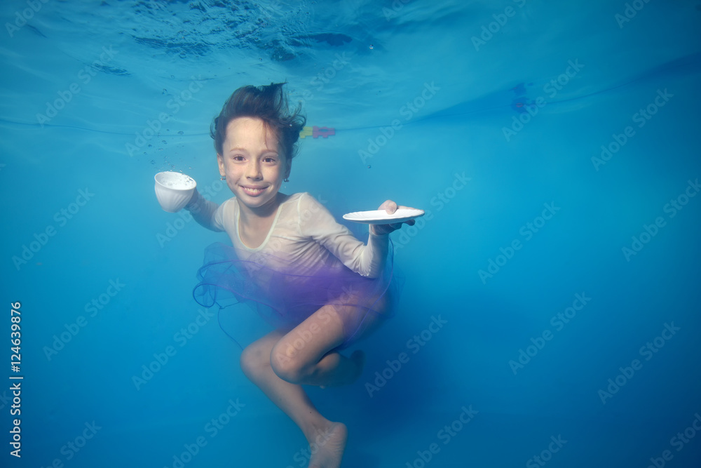 Happy little girl swimming underwater in the pool, posing with a white Cup and saucer in his hands and smiling. Portrait. Horizontal view