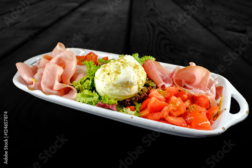 Appetizer plate with prosciutto crudo, chopped tomatoes, salad and burrata. Studio shot on black wooden background.