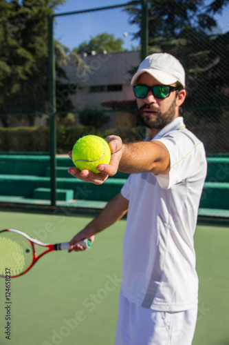 Professional tennis player, bearded young man offering tennis ball to play the game. He has a racket in her right hand and wears a white cap.  © pablobenii