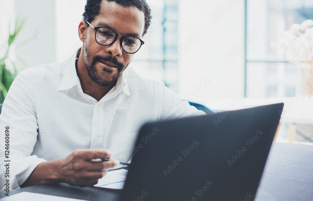Dark skinned coworker wearing glasses and using laptop in modern office.African american man in white shirt working on workplace.Horizontal,blurred background Dark skinned coworker wearing glasses and using laptop in modern office.African american man in white shirt working on workplace.Horizontal,blurred background