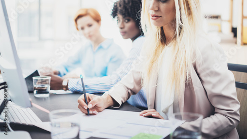 Beautiful blonde woman signing documnet on workplace in office. Group of girls coworkers discussing together business project. Horizontal, blurred background.