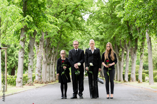 Familie läuft auf Allee im Friedhof in Trauer mit Blumen in der Hand