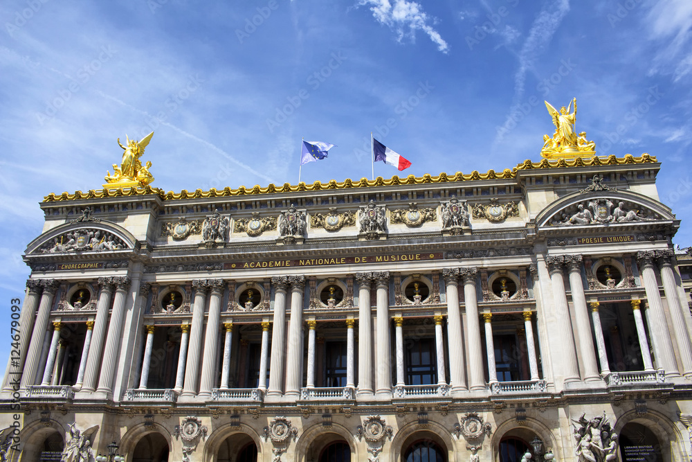 Bottom view of historical, old Paris opera house with clear blue sky background. European and French flags are waving on the top of the building. Golden statues are also in the view.