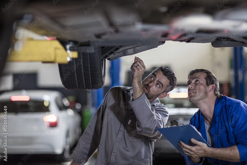 Two car mechanics in a workshop examining car together Two car mechanics in a workshop examining car together