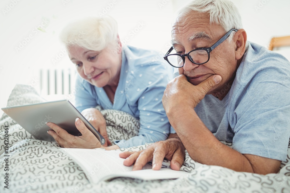 Senior couple reading a book and using digital tablet on bed