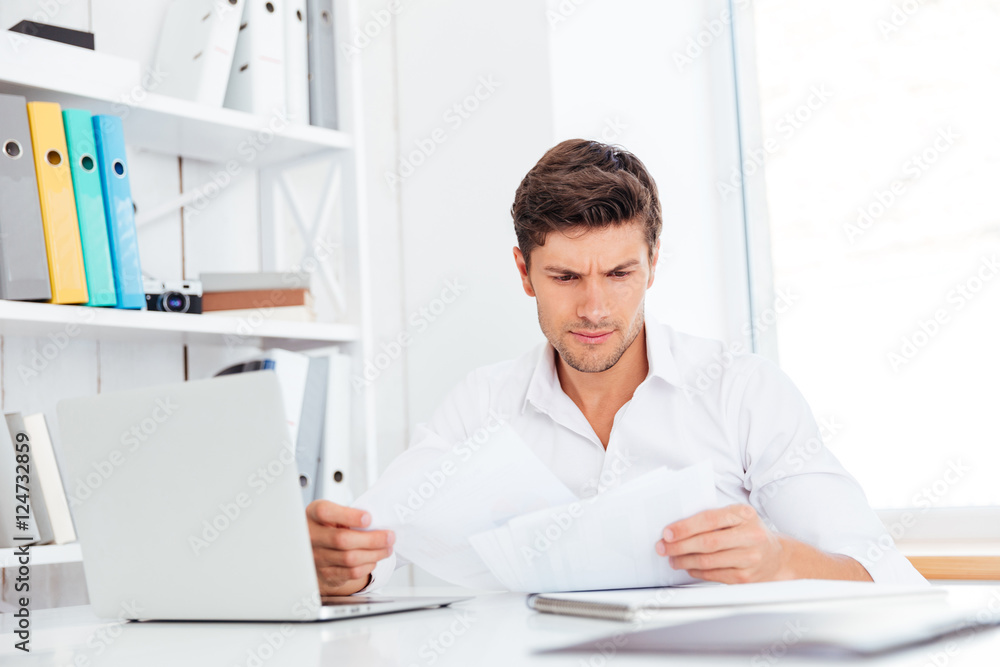 Handsome young businessman analyzing documents and laptop in office