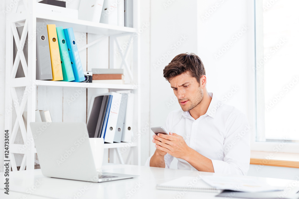 Pensive young businessman sitting at the table and using smartphone Pensive young businessman sitting at the table and using smartphone