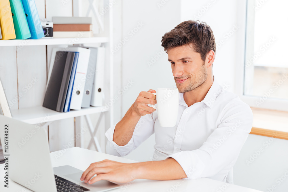 Young smiling businessman drinking coffee and typing on laptop Young smiling businessman drinking coffee and typing on laptop