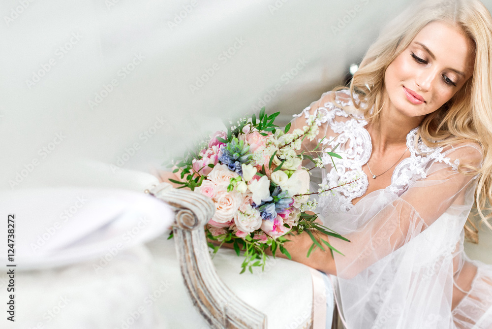 Naklejka premium Bride's morning. Fine art wedding. Portrait of a young bride in white lace boudoir with wavy blonde hair and a bouquet in her hands posing looking down shyly