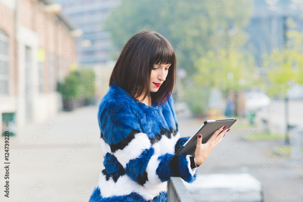 Knee figure of young beautiful eastern brown hair woman leaning against a wall, looking down and tapping the screen of a tablet hand hold - technology, social network, business concept Knee figure of young beautiful eastern brown hair woman leaning against a wall, looking down and tapping the screen of a tablet hand hold - technology, social network, business concept