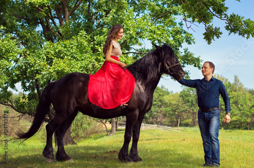 Young beautiful european woman in red dress sitting on a black horse and man standing nearby. Dating in the park. Love, family and relationship concept