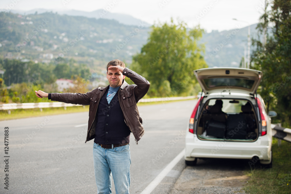 Hitchhiking - Need a drive. Young man on the road with his hand raised in front of car Hitchhiking - Need a drive. Young man on the road with his hand raised in front of car