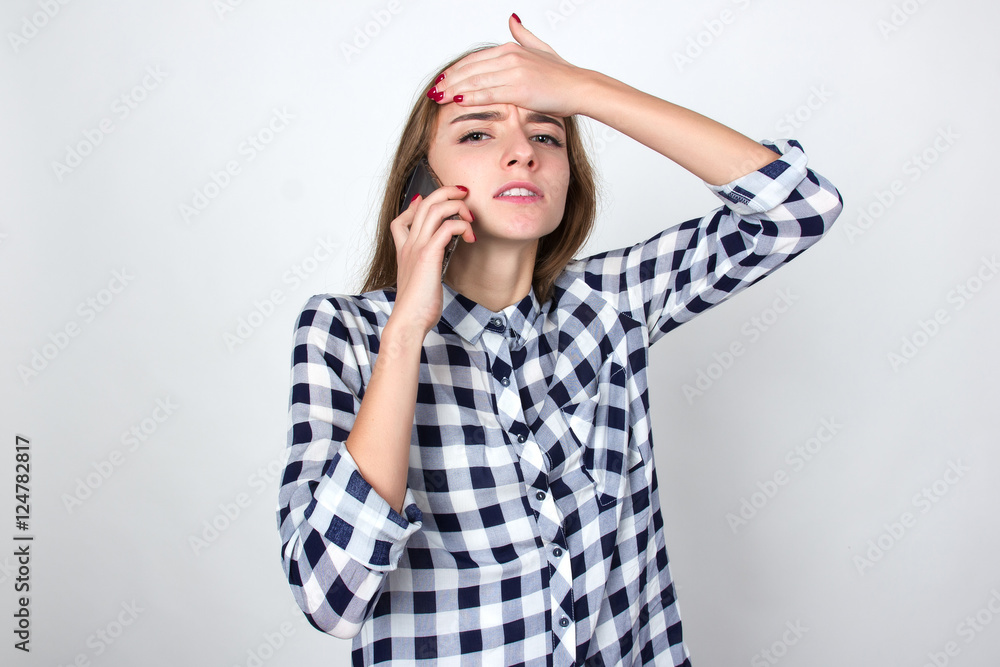 Portrait of a pretty woman in plaid shirt stress and headache having migraine pain and she calling a doctor standing over gray background and looking at camera Portrait of a pretty woman in plaid shirt stress and headache having migraine pain and she calling a doctor standing over gray background and looking at camera