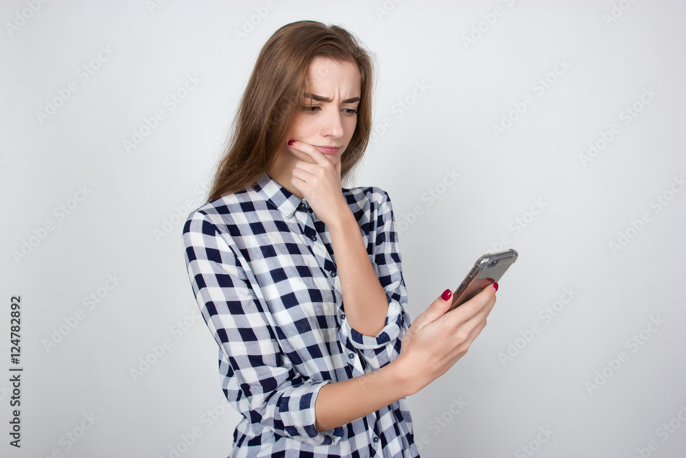 Portrait of a pretty thinking woman in plaid shirt standing over gray background and looking at phone Portrait of a pretty thinking woman in plaid shirt standing over gray background and looking at phone