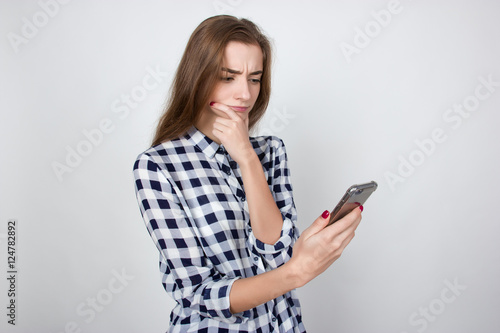 Portrait of a pretty thinking woman in plaid shirt standing over gray background and looking at phone