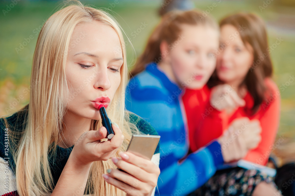 Young woman being gossiped about. Young beautiful blond woman doing make up while her friends communicating on background. Young woman being gossiped about. Young beautiful blond woman doing make up while her friends communicating on background.