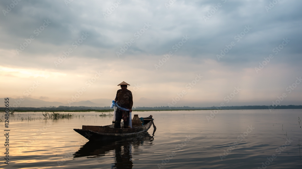 Asian fisherman on wooden boat casting a net for catching freshwater fish in nature river in the early morning before sunrise